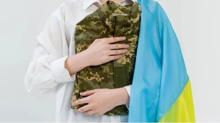 Жінка в білій сорочці тримає камуфляжний військовий мундир і український прапор